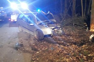 Zdjęcie przedstawia forda fokusa z uszkodzonym przodem pojazdu. Jego kierowca uderzył w drzewo. W tle widać niebieskie światła błyskowe wozu strażackiego