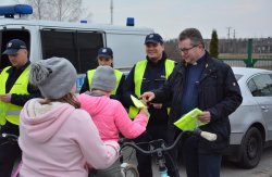 Policjanci z Komisariatu Policji w Sompolnie, Wydziału Prewencji, Wydziału Ruchu Drogowego oraz kapelan konińskich policjantów wręczają grupie dzieci na rowerach kamizelki i opaski odblaskowe oraz lampki rowerowe